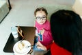 Cute little girl in eyeglasses using smartphone looking at her mother indoor in lobby or coffee shop. Children and