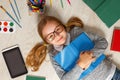 Cute little girl in glasses with a book lying on the floor. A child is surrounded by a book, tablet, paints, brushes, pencils Royalty Free Stock Photo