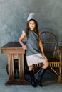 Cute little girl in grey dress smiling on camera with long hair stands next to a wooden chair and a table Royalty Free Stock Photo
