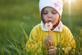 Cute little girl holding dandelion in hands and blowing, posing in green meadow, wearing yellow jacket and cap, female child Royalty Free Stock Photo