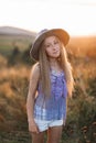 Cute little girl with long blond hair, wearing brown hat, posing in the wild flower and grass field with sunset Royalty Free Stock Photo