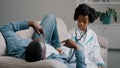 Cute little girl in medical gown plays with dad pretending to be doctor man lying on sofa funny daughter listening to Royalty Free Stock Photo