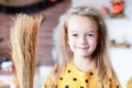 Cute little girl with messy hair, dressed up as a witch and holding a broom is standing in Halloween decorated living room,smiling