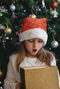 A cute little girl opens a Christmas present with surprise in a New Year`s hat under the tree. Holiday, present, miracle, magic Royalty Free Stock Photo