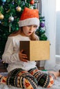 A cute little girl opens a Christmas present with surprise in a New Year`s hat under the tree. Holiday, present, miracle, magic Royalty Free Stock Photo