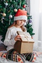 A cute little girl opens a Christmas present with surprise in a New Year`s hat under the tree. Holiday, present, miracle, magic. Royalty Free Stock Photo