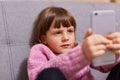 Cute little girl with pigtails wearing warm pink sweater, playing video game, using modern smartphone, smart small kid browsing Royalty Free Stock Photo