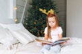 Cute little girl in pink dress reading book sitting on bed in room with Christmas tree. Child in rim with deer horns in cottage Royalty Free Stock Photo