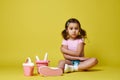 A cute little girl with ponytails sitting in summer clothes with beach toys: a bucket, a watering can and a rake with a shovel Royalty Free Stock Photo
