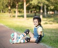 Cute little girl in protective equipment and rollers sitting on walkway in park outdoor. Summer activity for children