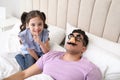 Cute little girl putting funny glasses on father while he sleeping in bed at home Royalty Free Stock Photo