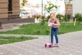 Cute little girl riding scooter in city park on bright summer day. Blond toddler having fun on street. Active leisure Royalty Free Stock Photo