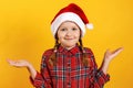Cute little girl in santa hat shrugs. Close-up of a child waiting for Christmas on a yellow background Royalty Free Stock Photo