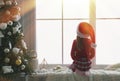 Cute little girl in Santa hat on window sill near Christmas tree at home, back view Royalty Free Stock Photo