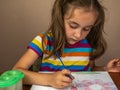 A cute little girl sits at a table and draws with paints on white paper. A beautiful little girl draws with pleasure Royalty Free Stock Photo