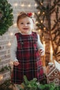 Cute little girl is sitting by the Christmas tree on the floor in the room. Near the baby is a gingerbread house and a basket with