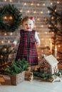 Cute little girl is sitting by the Christmas tree on the floor in the room. Near the baby is a gingerbread house and a basket with