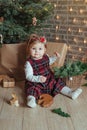 Cute little girl is sitting by the Christmas tree on the floor in the room. Near the baby is a gingerbread house and a basket with