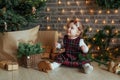 Cute little girl is sitting by the Christmas tree on the floor in the room. Near the baby is a gingerbread house and a basket with