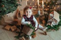 Cute little girl is sitting by the Christmas tree on the floor in the room. Near the baby is a gingerbread house and a basket with