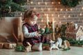 Cute little girl is sitting by the Christmas tree on the floor in the room. Near the baby is a gingerbread house and a basket with