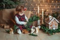 Cute little girl is sitting by the Christmas tree on the floor in the room. Near the baby is a gingerbread house and a basket with