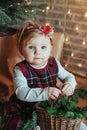 Cute little girl is sitting by the Christmas tree on the floor in the room. Near the baby is a gingerbread house and a basket with