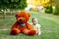 Cute little girl sitting on green grass with big Teddy bear in yellow summer dress in summer Royalty Free Stock Photo