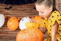 Cute little girl sitting on kitchen table carving large pumpkin. Halloween background.