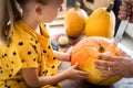 Cute little girl sitting on kitchen table, helping her father to carve large pumpkin, smiling. Halloween family lifestyle.