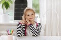 A cute little girl is sitting at a table with a sketchbook and a glass of colored pencils. The girl shows her heart with Royalty Free Stock Photo