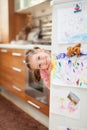 Cute little girl smiling behind refrigerator door in kitchen