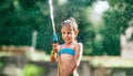 Cute little girl sprinkling with water for herself from the hose, making a rain pleasure for hot summer days. Careless childhood