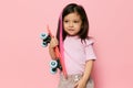 a cute little girl is standing on a pink background in a pink T-shirt, holding her skate in her hands, looking at it Royalty Free Stock Photo