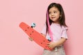 a cute little girl is standing on a pink background in a pink T-shirt, holding her skate in her hands, looking at it Royalty Free Stock Photo