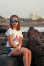 Cute little girl in sunglasses sitting on a rock against the backdrop of the ocean. Family seaside vacation. Royalty Free Stock Photo