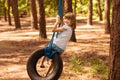 Cute little girl swinging on wheel attached to big tree in forest.
