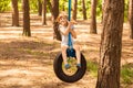 Cute little girl swinging on wheel attached to big tree in forest.