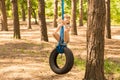 Cute little girl swinging on wheel attached to big tree in forest.