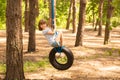 Cute little girl swinging on wheel attached to big tree in forest.