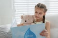 Cute little girl with teddy bear reading book on sofa at home Royalty Free Stock Photo