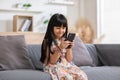 Cute little girl uses smartphone while sitting at the sofa in the living room. Child surfing the internet on mobile phone, browses Royalty Free Stock Photo