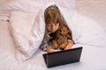 Cute little girl using a laptop in bed under a blanket Royalty Free Stock Photo