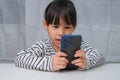 Cute little girl using smartphone and smiling while sitting at table in house. Happy Asian girl holding phone in hands, online Royalty Free Stock Photo