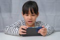 Cute little girl using smartphone and smiling while sitting at table in house. Happy Asian girl holding phone in hands, online Royalty Free Stock Photo