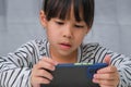 Cute little girl using smartphone and smiling while sitting at table in house. Happy Asian girl holding phone in hands, online Royalty Free Stock Photo