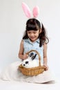 Cute little girl wearing pink bunny ears and holding rabbit in basket while sitting on white background. Joyful kid celebrating Royalty Free Stock Photo