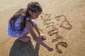 Cute little girl wearing a white dress is writting a phrase `I Love Summer` on the sand Royalty Free Stock Photo