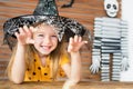 Cute little girl wearing witch hat sitting behind a table in Halloween theme decorated living room, making scary face. Royalty Free Stock Photo