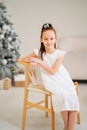 Cute little girl in white dress sitting on a chair by Christmas tree. Royalty Free Stock Photo